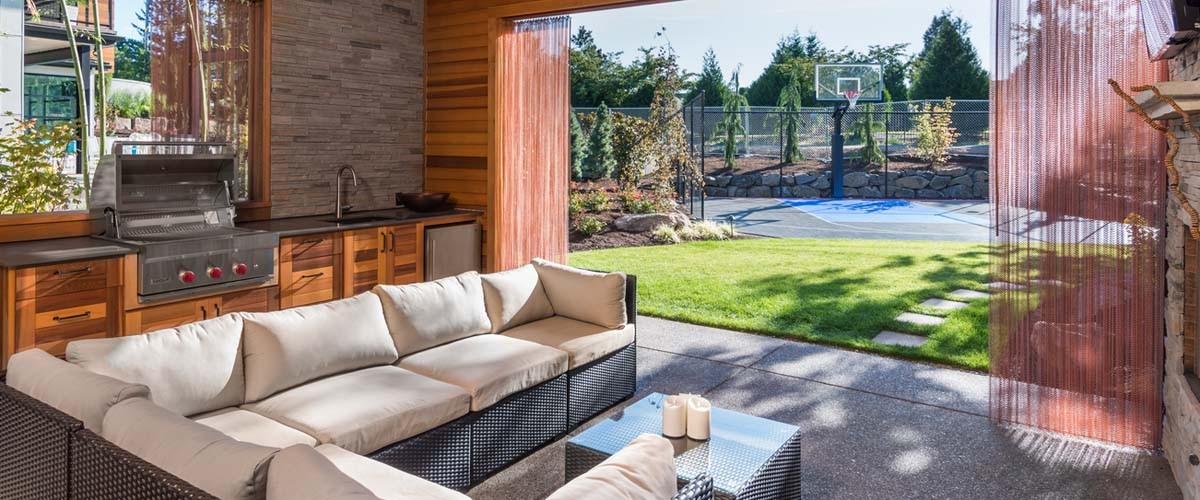Covered patio behind new luxury home with fireplace, tv, and couch, with view of lush green grass and landscaping